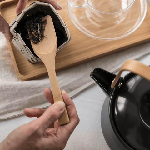 A person uses a bamboo scoop to scoop up loose leaf tea. A teapot, tea cup and tray are nearby.