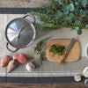 A bamboo Bar Board is ready for cooking prep. Herbs are on and around the board, along with potatoes and a head of garlic. A stockpot waits for all ingredients to be combined.