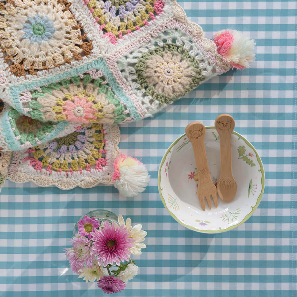 Organic Bamboo Kid's Fork & Spoon are resting in a child's bowl atop a blue checkered tablecloth. A colorful crocheted baby blanket is near, along with a small bouquet of pink and white flowers.