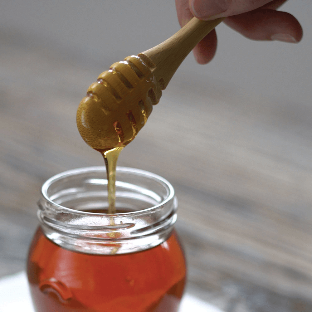 An Organic Bamboo Honey Dipper that was just dipped into a jar of honey is held, with excess honey dripping back into the jar.