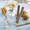 A set of Reusable Short Bamboo Straws are displayed in juice glasses on a table set with a blue checkered tablecloth - bambu