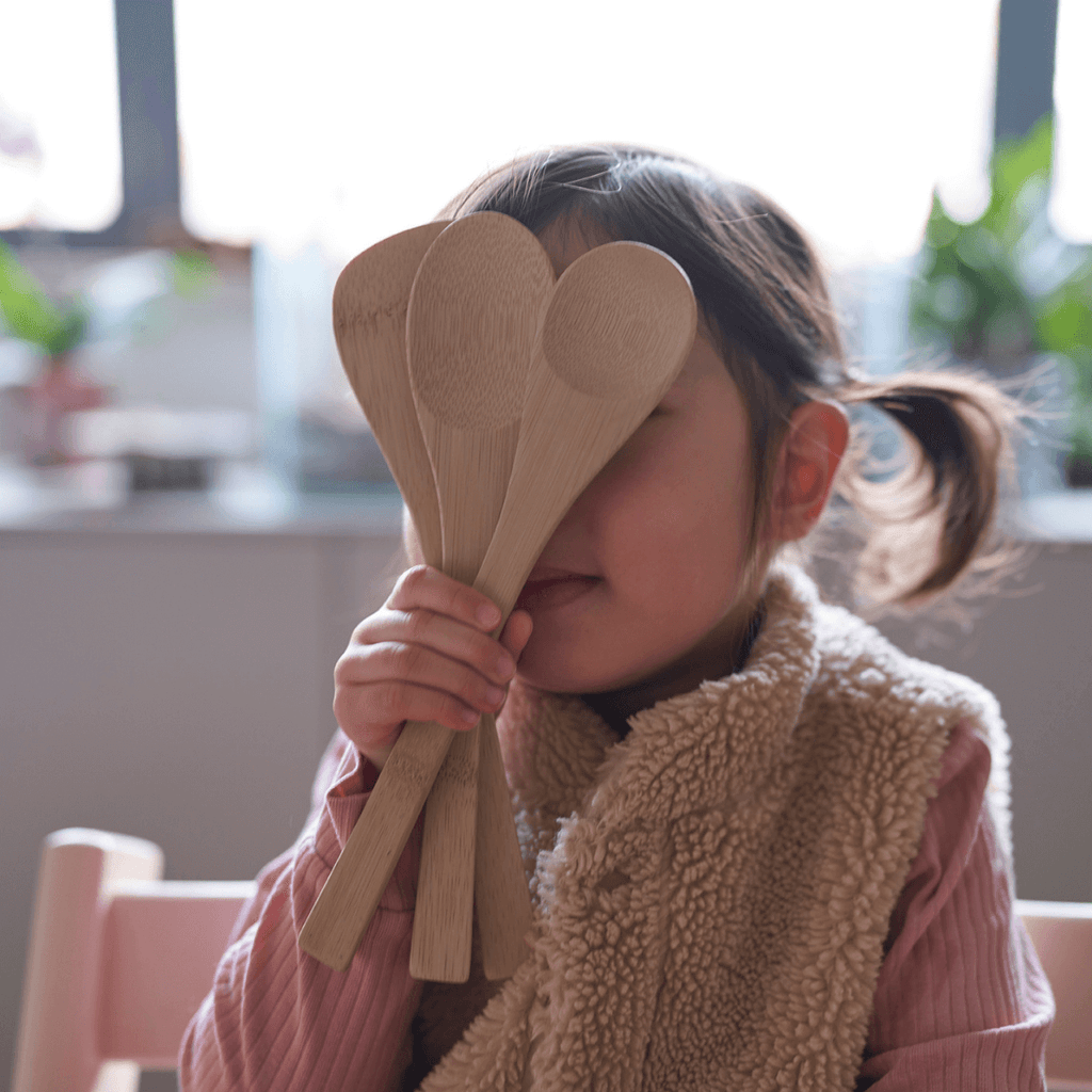 A young child holds a set of Kids in the Kitchen utensils in front of their face. bambu