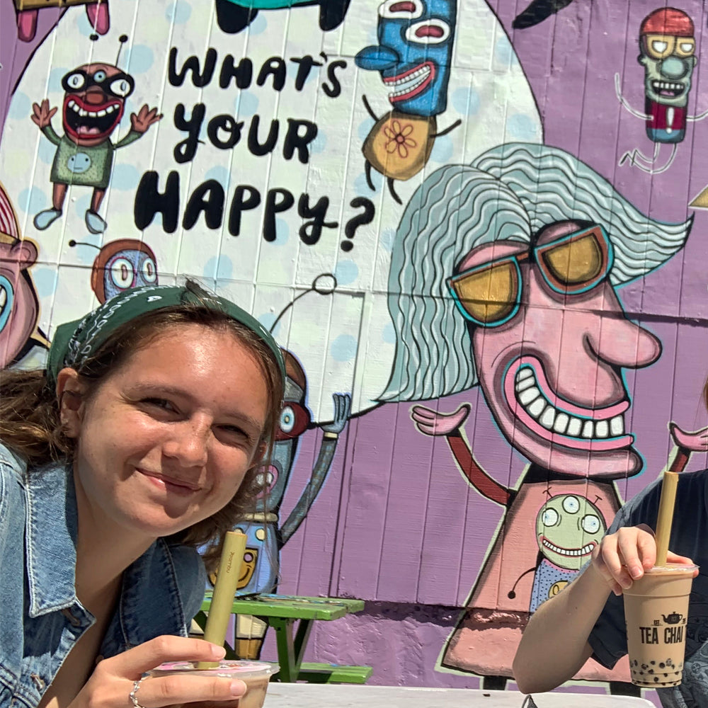 A young woman sits in front of a colorful mural. She holds a Boba Tea with a Reusable Bamboo Jumbo Straw.