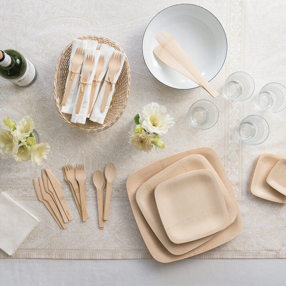 A stack of Veneerware® Square Bamboo Plates in various sizes is on a table. Veneerware citlery, drinking glasses, and a serving bowl are nearby.