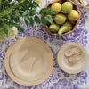 Veneerware® Round Bamboo Plates are stacked on a table that is covered with a purple flower tablecloth. There is a bowl of pears and a vase of flowers on the table.