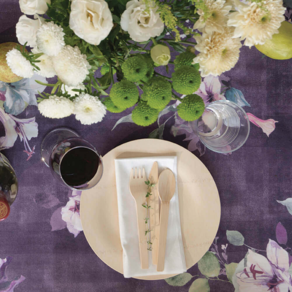 A 9 inch Veneerware® Round Bamboo Plate is on a purple tablecloth. There is a bamboo napkin on top of the plate with a set of Veneerware cutlery.