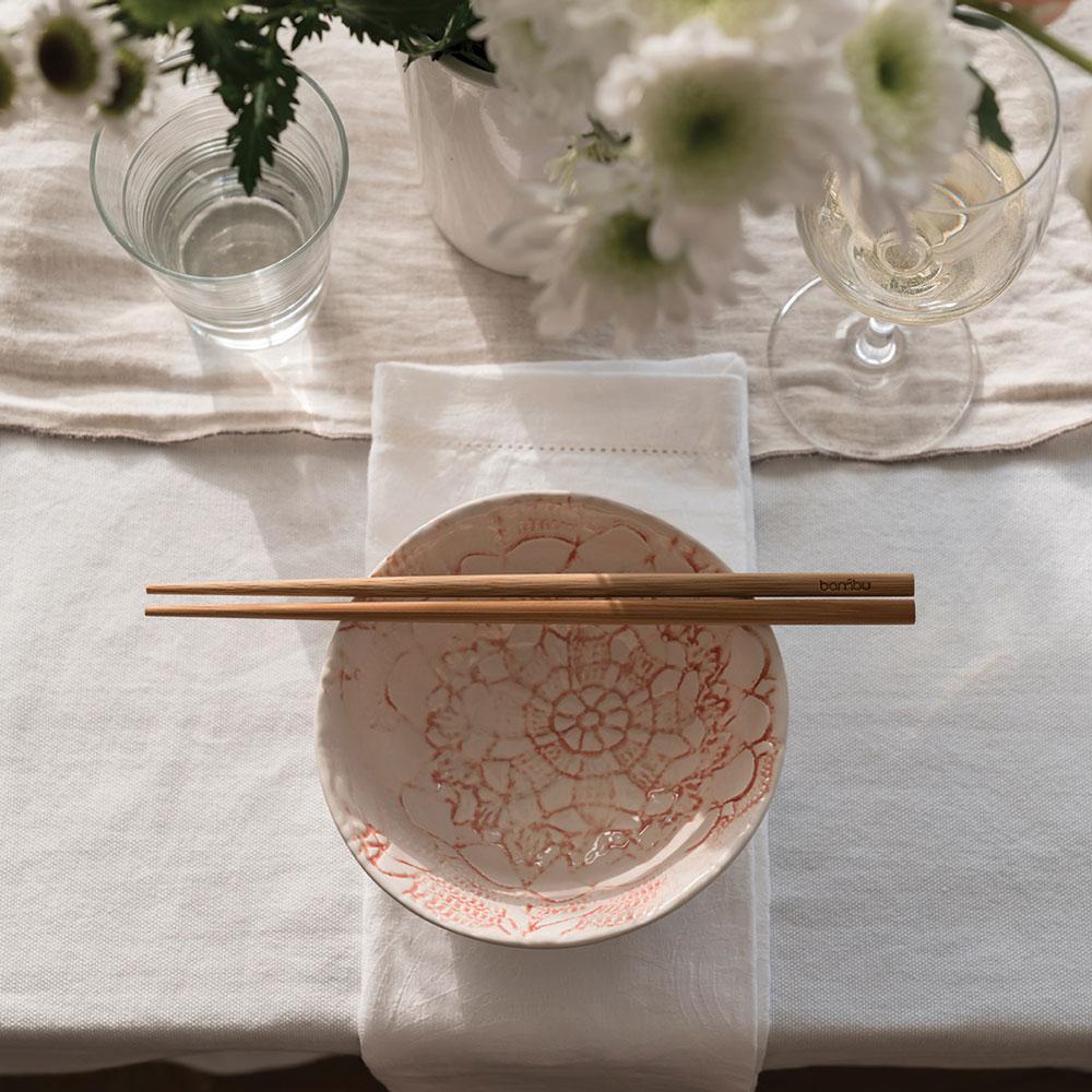 A pair of Reusable Bamboo Chopsticks are laid across a blue and pink patterned ceramic bowl. A cloth napkin is under the bowl. A water glass and wine glass are placed nearby.