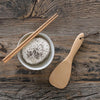 A set of Reusable Bamboo Chopsticks lay across a bowl of rice. A bamboo rice paddle is on the tabletop next to the bowl.
