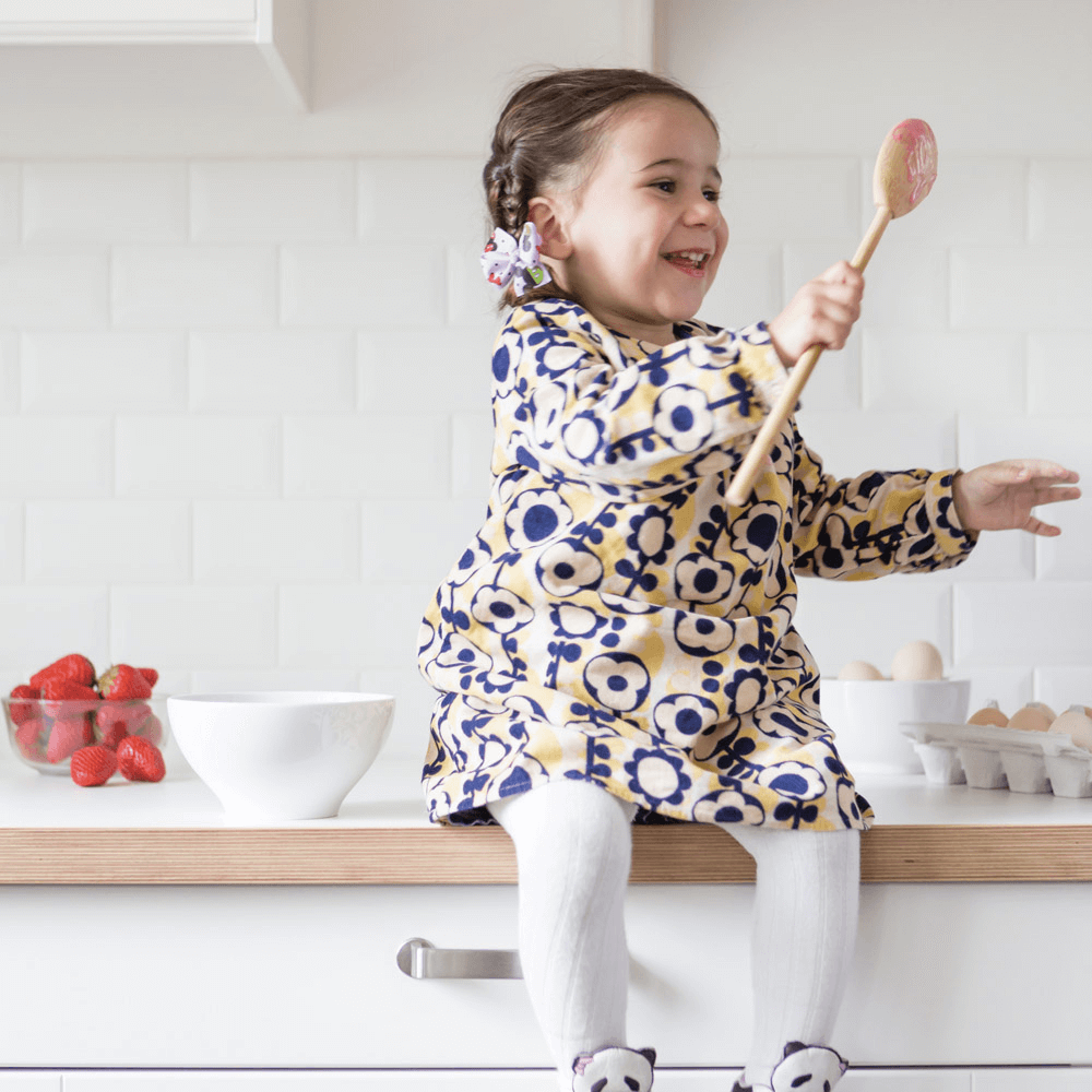 A child in a floral dress is sitting on a kitchen counter, holding a bamboo mixing spoon, There is pink batter on the spoon and a bowl of strawberries is on the counter.